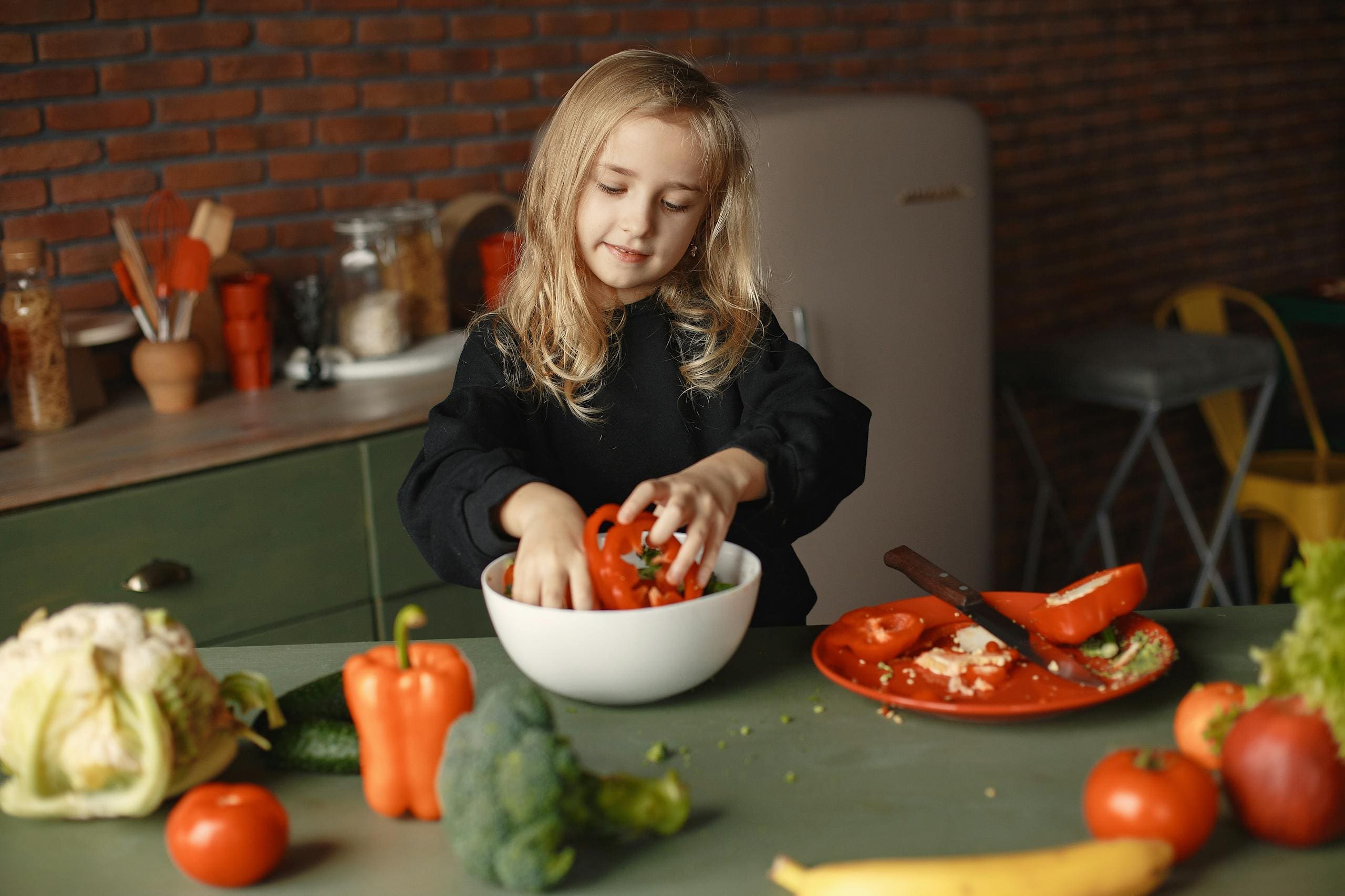 A young girl in the kitchen preparing a healthy vegetable salad with fresh ingredients featuring peppers, broccoli, and tomatoes.