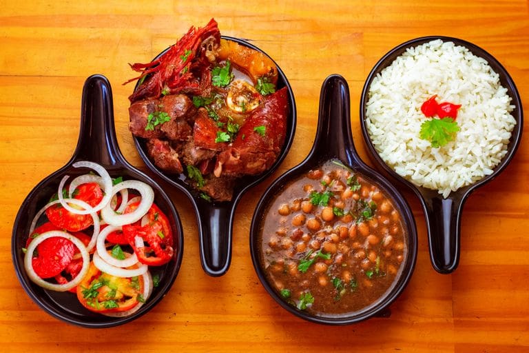 Top view of traditional Brazilian dishes with rice, beans, beef, and fresh salad served in black bowls.